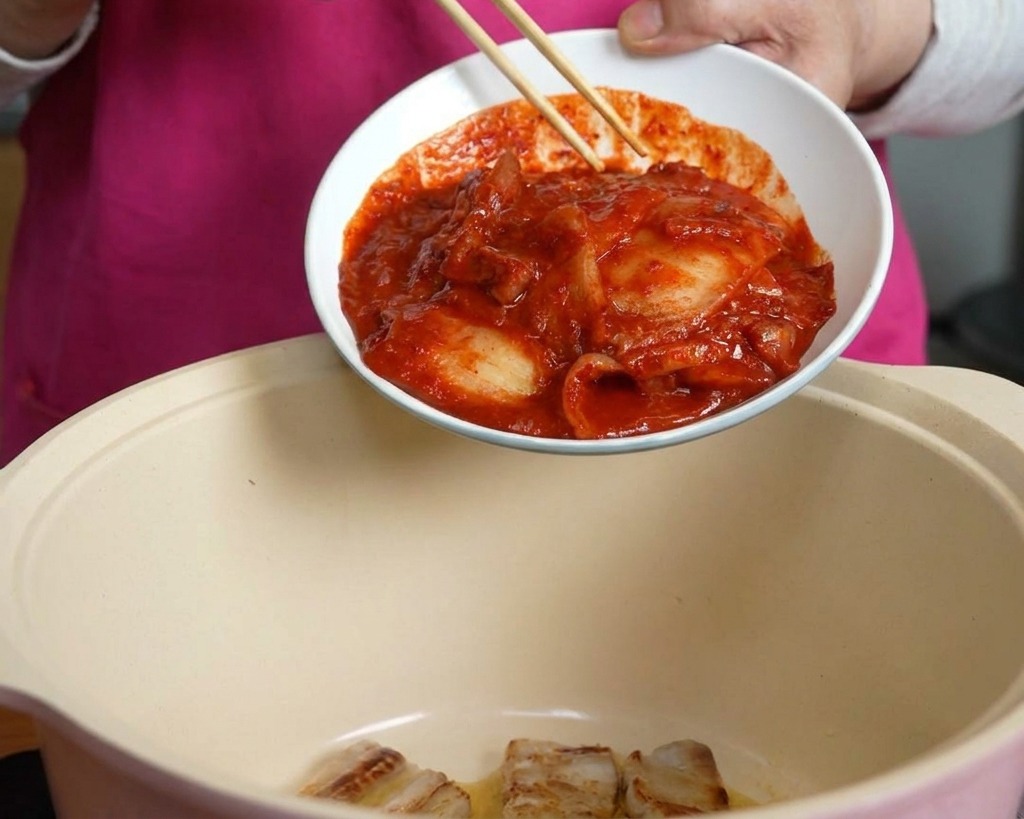 Dłoń za pomocą pałeczek zsuwa miskę pikantnego, czerwonego kimchi do garnka z boczkiem wieprzowym.