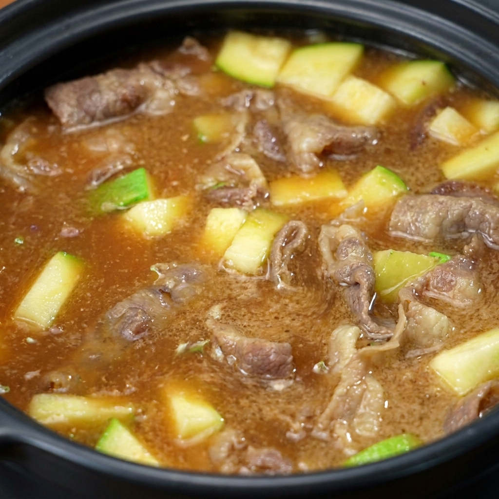 Pasta sojowa mieszająca się z wodą w garnku, zmieniająca płyn w mętny, brązowy bulion z widocznymi kawałkami wołowiny i cukinii.