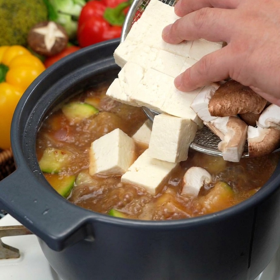 Dłoń delikatnie przekładająca kostki białego tofu i plastry brązowych grzybów shiitake do gotującej się zupy sojowej.