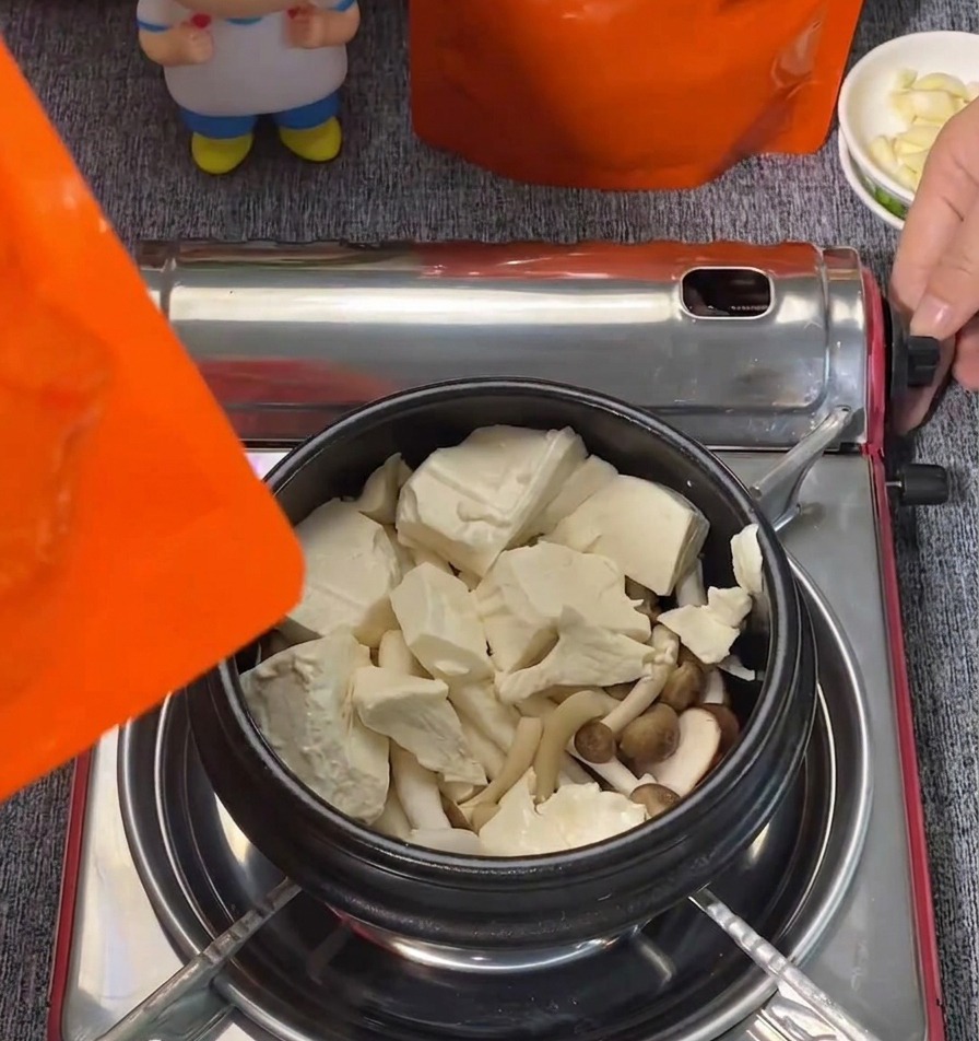 Duże bloki białego miękkiego tofu i grzyby shimeji ułożone w czarnym garnku.