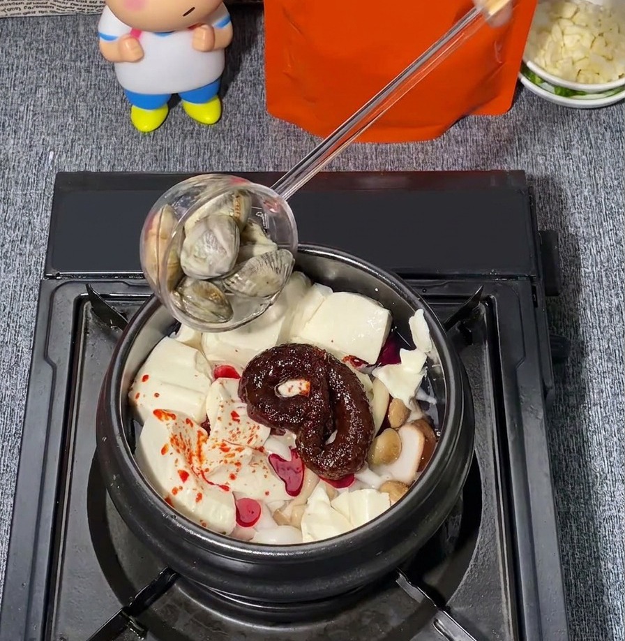 Świeże małże dodawane chochlą do kamiennego garnka zawierającego miękkie tofu, grzyby i czerwoną przyprawę chili.
