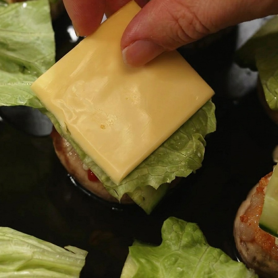 Dłoń kładąca kwadratowy plasterek żółtego sera na sałacie i ogórku wewnątrz burgera pieczarkowego.