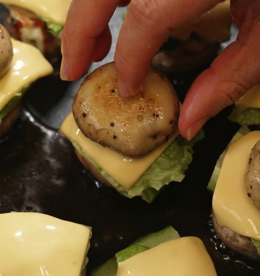 Dłoń nakładająca grillowany wcześniej kapelusz pieczarki na mini burgera, kończąc składanie dań na patelni.