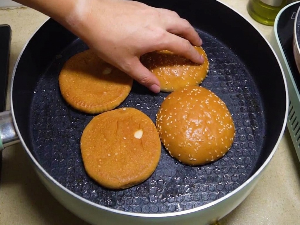Połówki bułek do burgera opiekanie wewnętrzną stroną do dołu na patelni, a dłoń poprawia ich ułożenie.