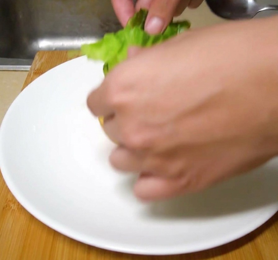 Świeże liście zielonej sałaty układane dłonią na opieczonej bułce do burgera na białym ceramicznym talerzu.