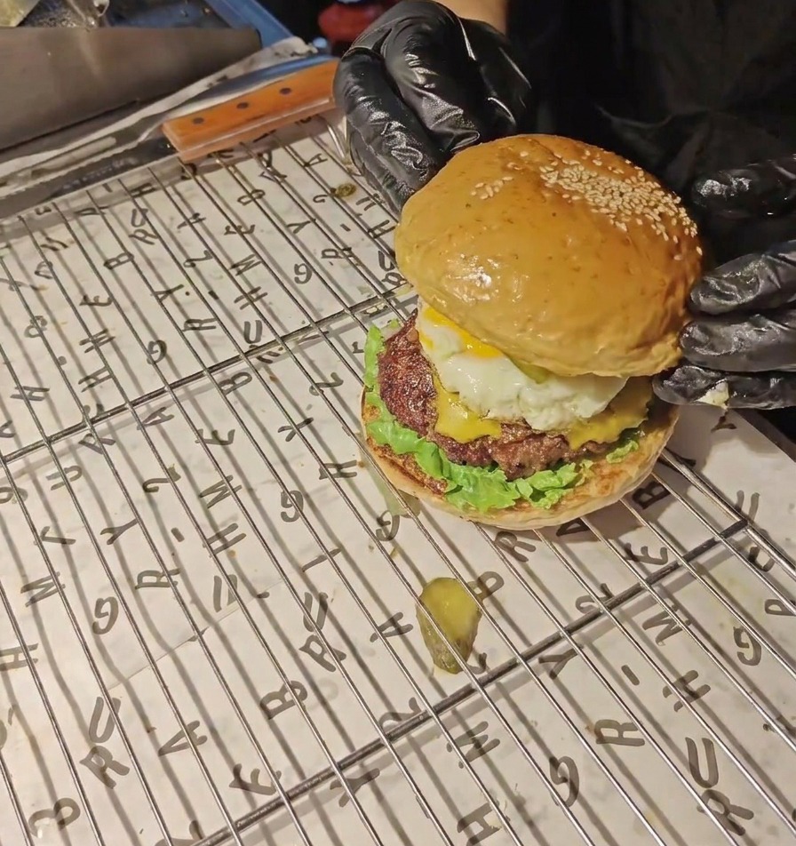 Dłonie w czarnych rękawiczkach kładą złocistą bułkę z sezamem na samym wierzchu gotowego burgera.