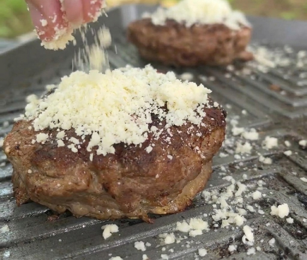 Zbliżenie na tarty ser sypany na zrumieniony kotlet wołowy na gorącej patelni grillowej.