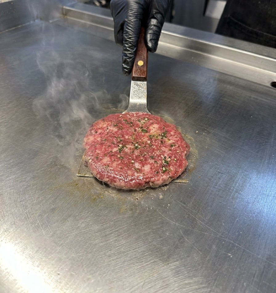 Metalowa łopatka dociska kotlet wołowy na stalowej płycie grillowej, z której unosi się para.
