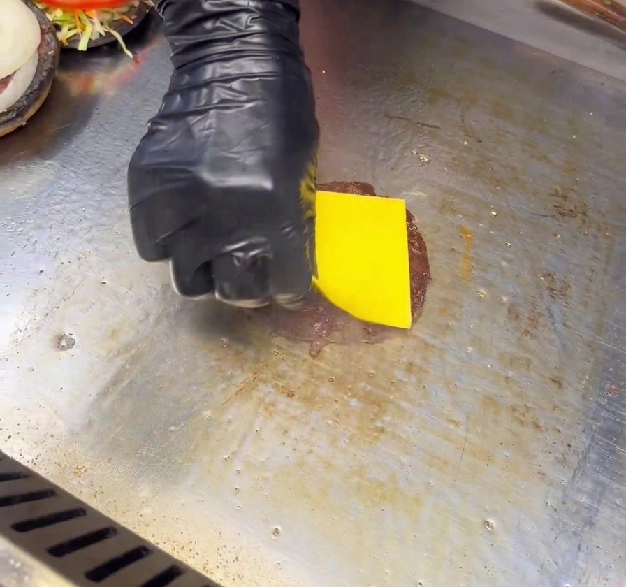 Dłoń w czarnej rękawiczce kładzie plaster żółtego sera na wysmażonym kotlecie burgerowym na płaskim grillu.