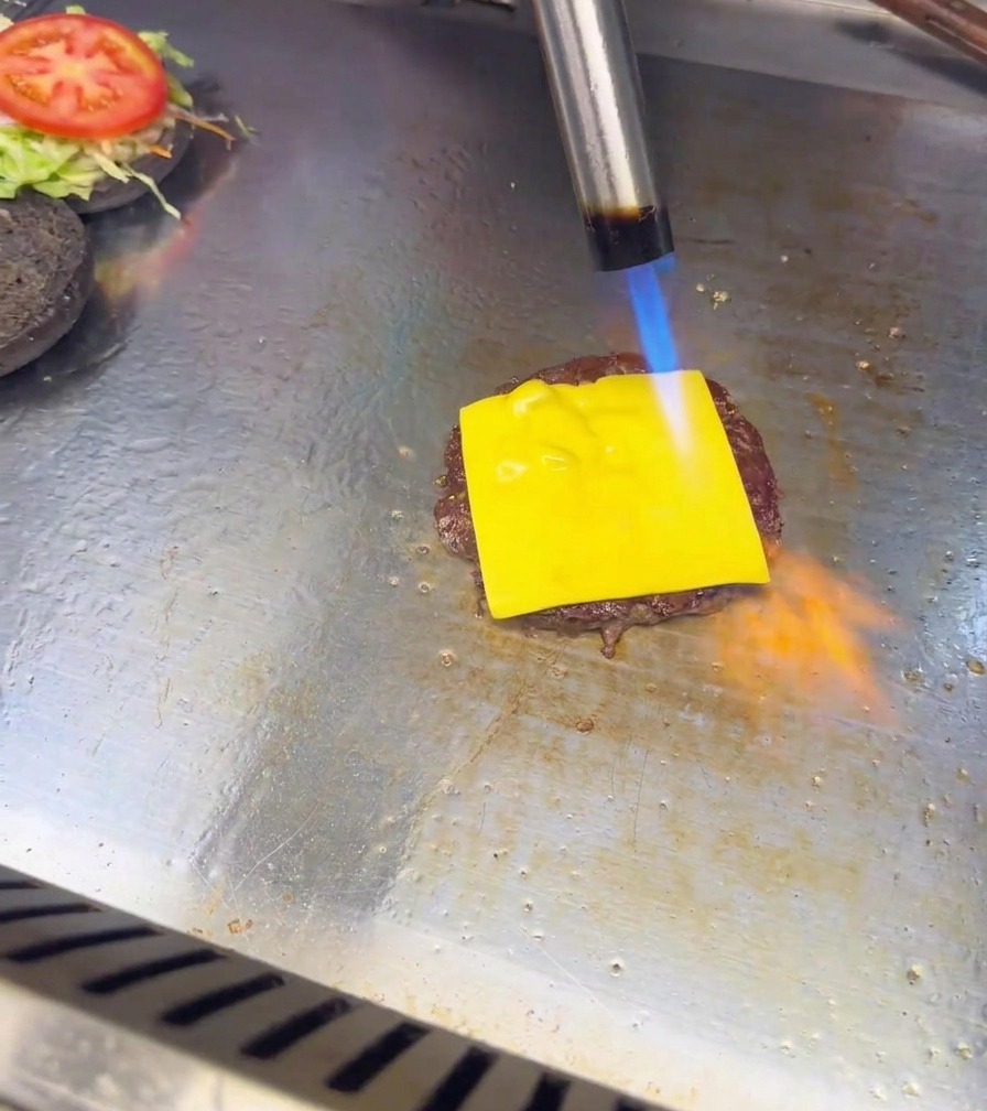 Niebiesko-pomarańczowy płomień z palnika kuchennego roztapia plaster sera na kotlecie burgerowym.