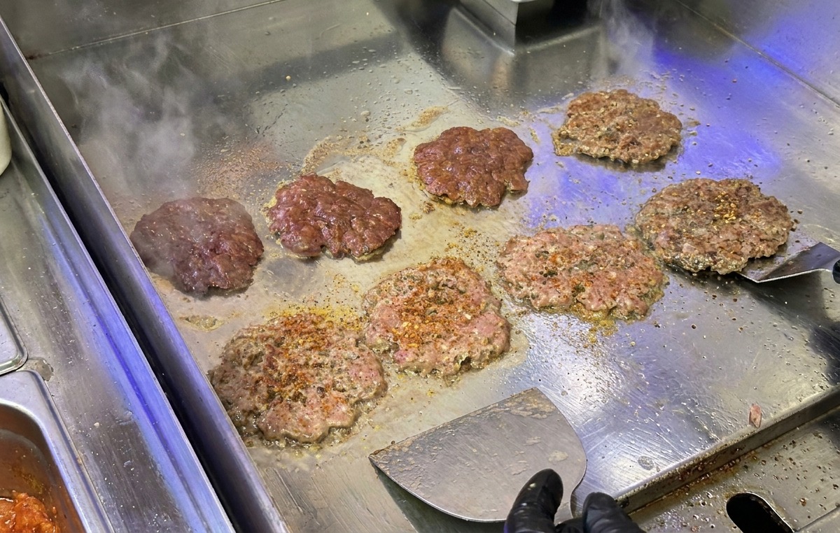 Kotlety wołowe do burgerów dociskane i smażone na stalowej płycie grillowej przy użyciu metalowej szpatułki.