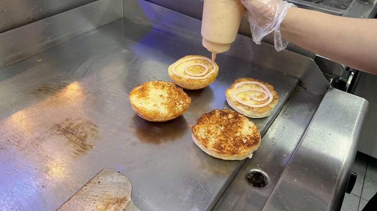 Kremowy różowy sos nakładany okrężnymi ruchami na opieczone bułki brioche na płaskim grillu.