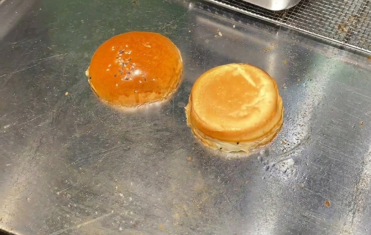 Dwie połówki bułki do burgera podpiekające się wnętrzem do dołu na płaskiej, metalowej powierzchni grilla.