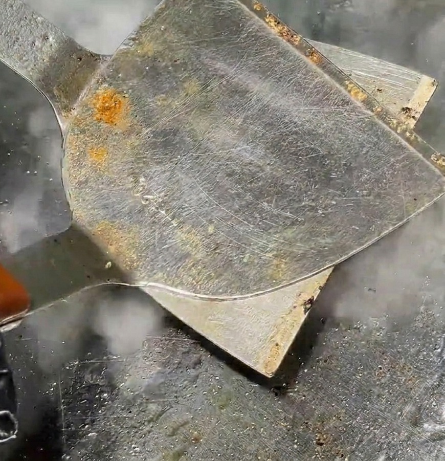 Metalowa łopatka dociskająca okrągłą kulę mięsa na płaski kotlet na gorącym grillu z unoszącą się parą.