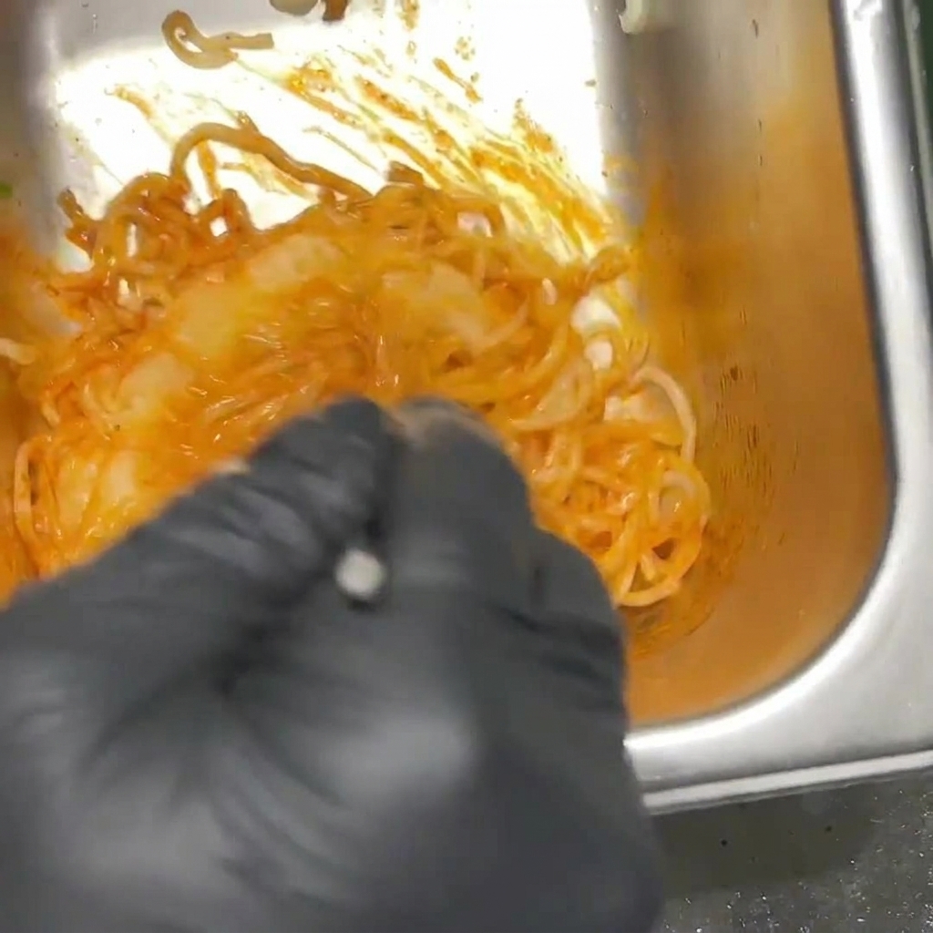Pikantny, pomarańczowy makaron ramen mieszany w metalowym pojemniku przez osobę w czarnych rękawiczkach.