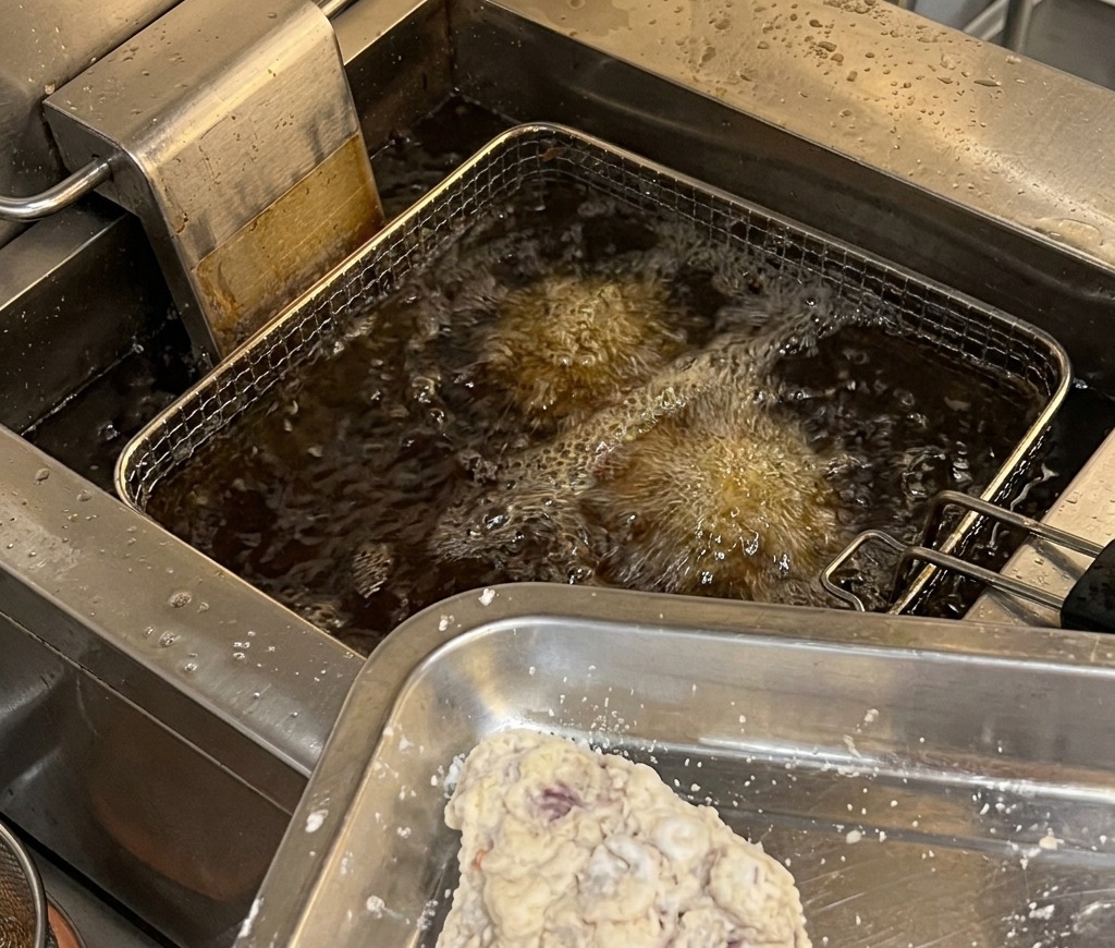 Opanierowane kawałki kurczaka na metalowej tacy przygotowane do włożenia do gastronomicznej frytkownicy z gorącym, bulgoczącym olejem.