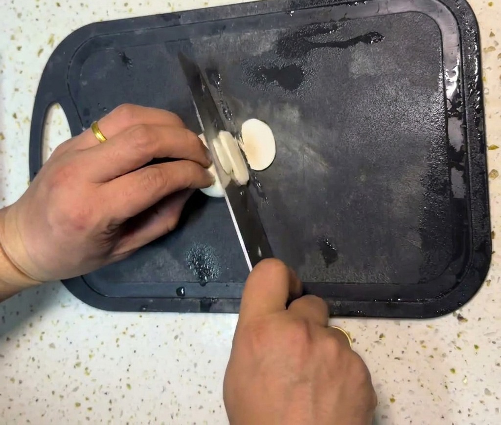 Ręce osoby krojącej białe pieczarki na cienkie plasterki za pomocą noża szefa kuchni na czarnej desce do krojenia.