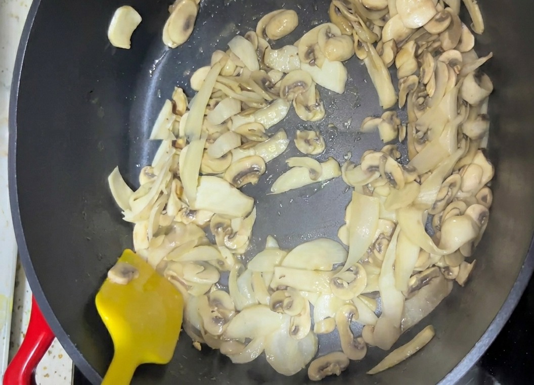 Pokrojone grzyby mieszane na ciemnej patelni za pomocą jasnożółtej łopatki.