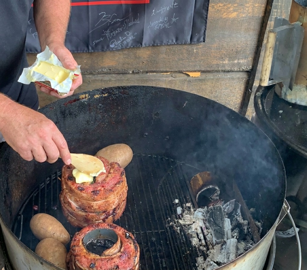 Drewniana łyżka nakładająca kawałek masła na mięso na grillu węglowym obok całych ziemniaków.