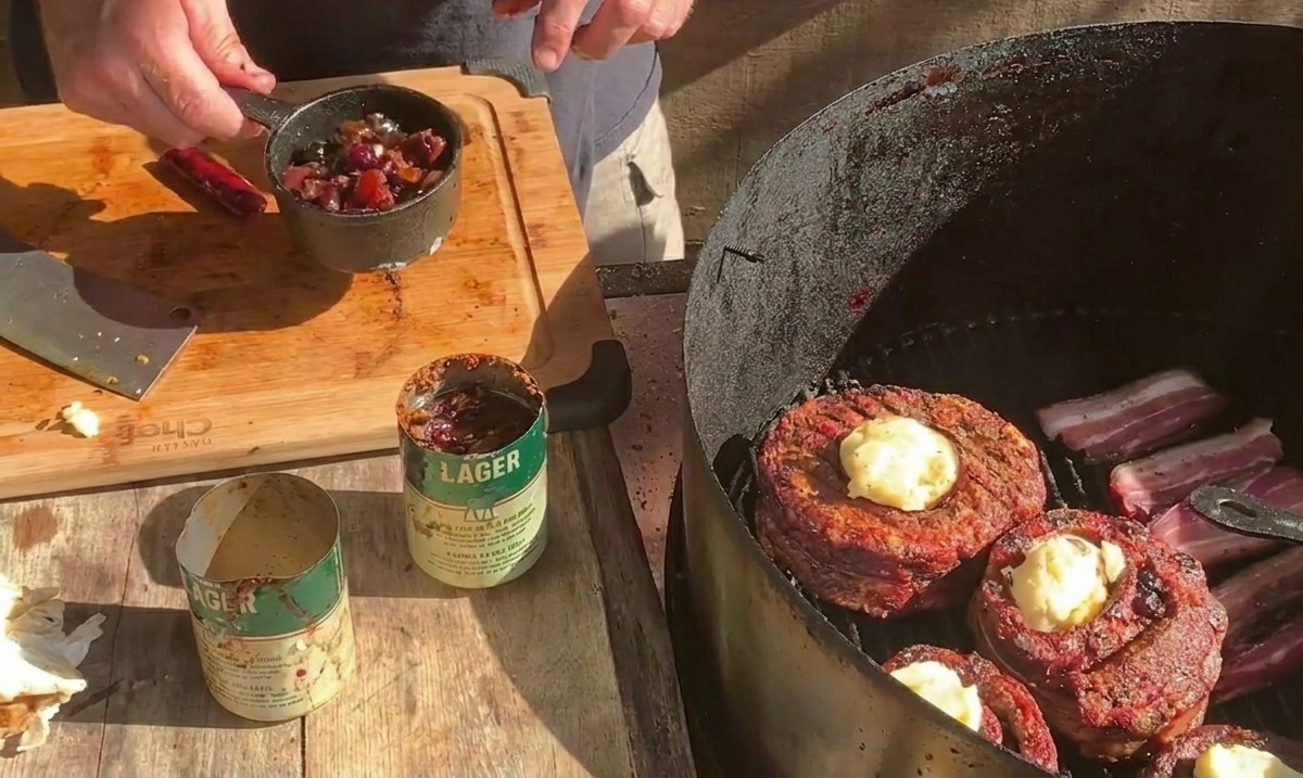 Osoba trzymająca mały metalowy garnuszek z sosem jagodowym nad grillem, gdzie koszyczki owinięte boczkiem są posypywane serem.