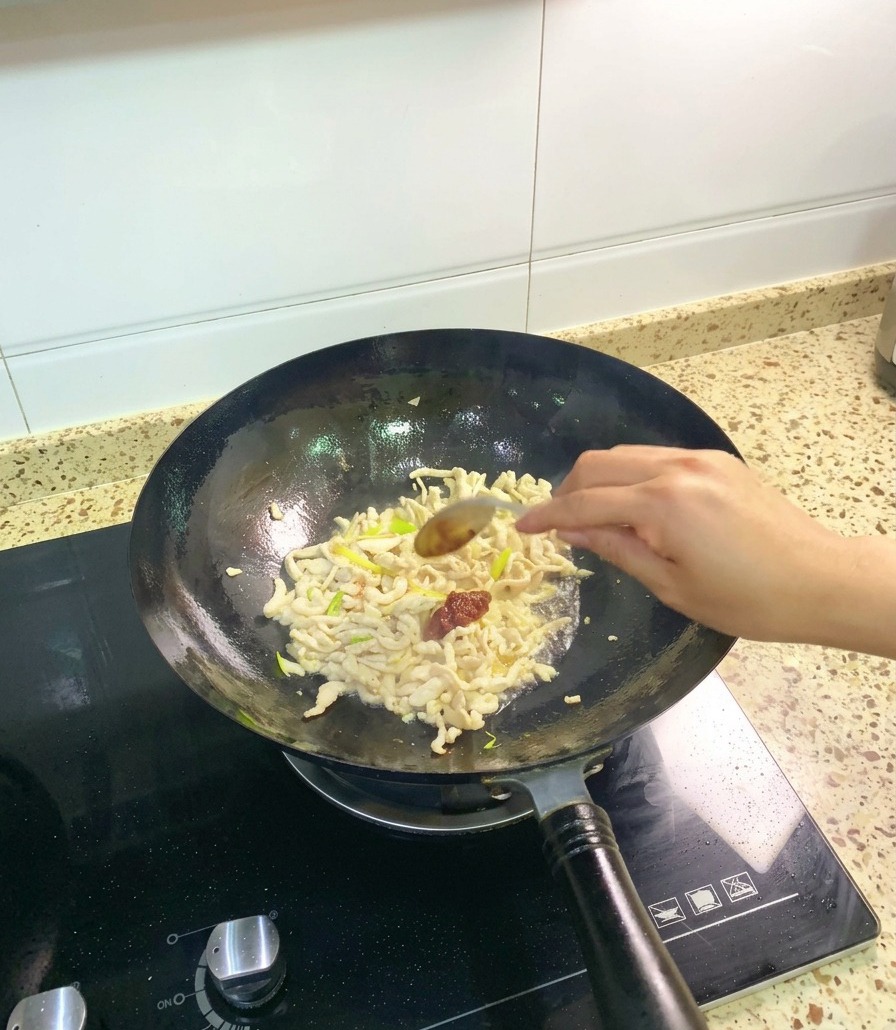 Stalowa łyżka nakładająca porcję gęstej, czerwonej pasty z bobu Pixian do woka z podsmażonym kurczakiem i dodatkami.