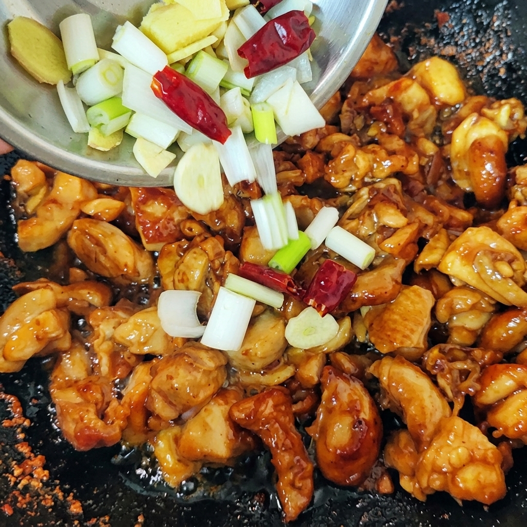 Mieszanka posiekanej zielonej dymki, plasterków imbiru i czerwonych suszonych papryczek chili wsypywana z małej miseczki do woka z podsmażonym kurczakiem.