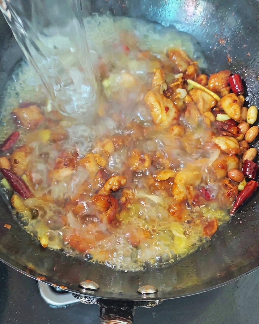 Strumień wrzącej wody wlewany do czarnego woka z podsmażonym kurczakiem i czerwonymi papryczkami chili, tworzący bulgoczący bulion.