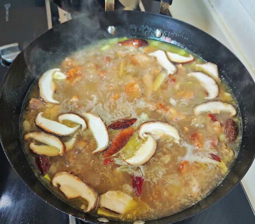 Pokrojone grzyby shiitake i czerwone papryczki chili gotujące się w bulgoczącym złocistym bulionie z kurczaka w czarnym woku.