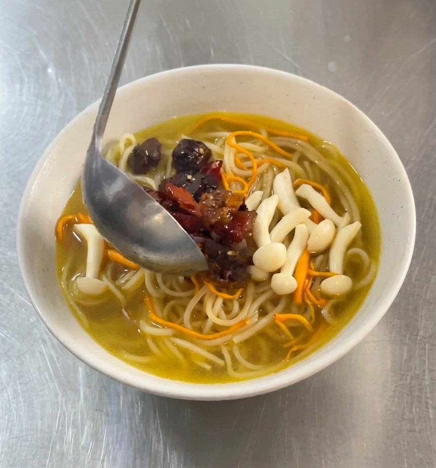 Metalowa łyżka nakładająca kawałki pikantnego kurczaka w czerwonej marynacie oraz suszone papryczki chili do miski z białym makaronem w złocistym rosole grzybowym.