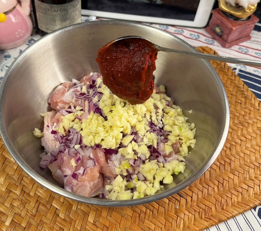 Metalowa łyżka z dużą porcją intensywnie czerwonej pasty Gochujang nad miską z surowym kurczakiem, czosnkiem i cebulą.