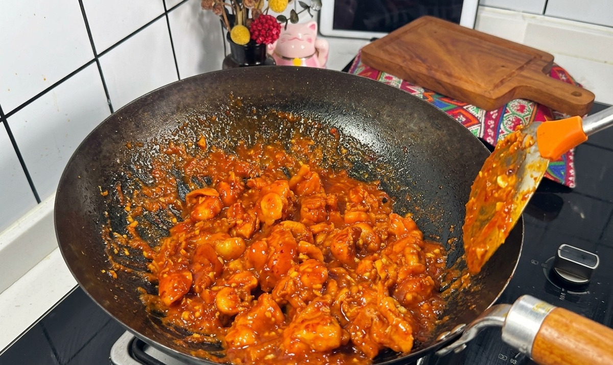 Kawałki kurczaka pokryte gęstym, bulgoczącym czerwonym sosem smażone w czarnym woku przy użyciu metalowej łopatki.