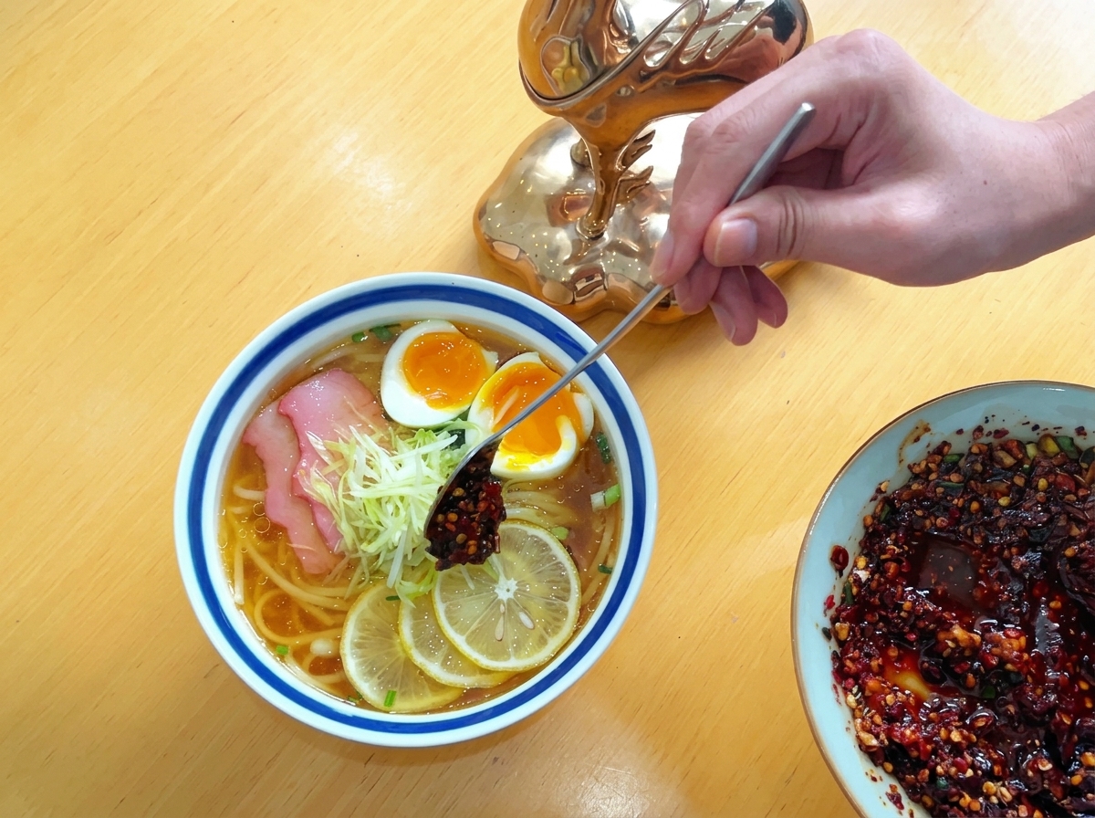 Osoba nakładająca długą metalową łyżką porcję ciemnoczerwonego oleju chili na kopczyk poszatkowanej białej rzodkwi w misce z makaronem.