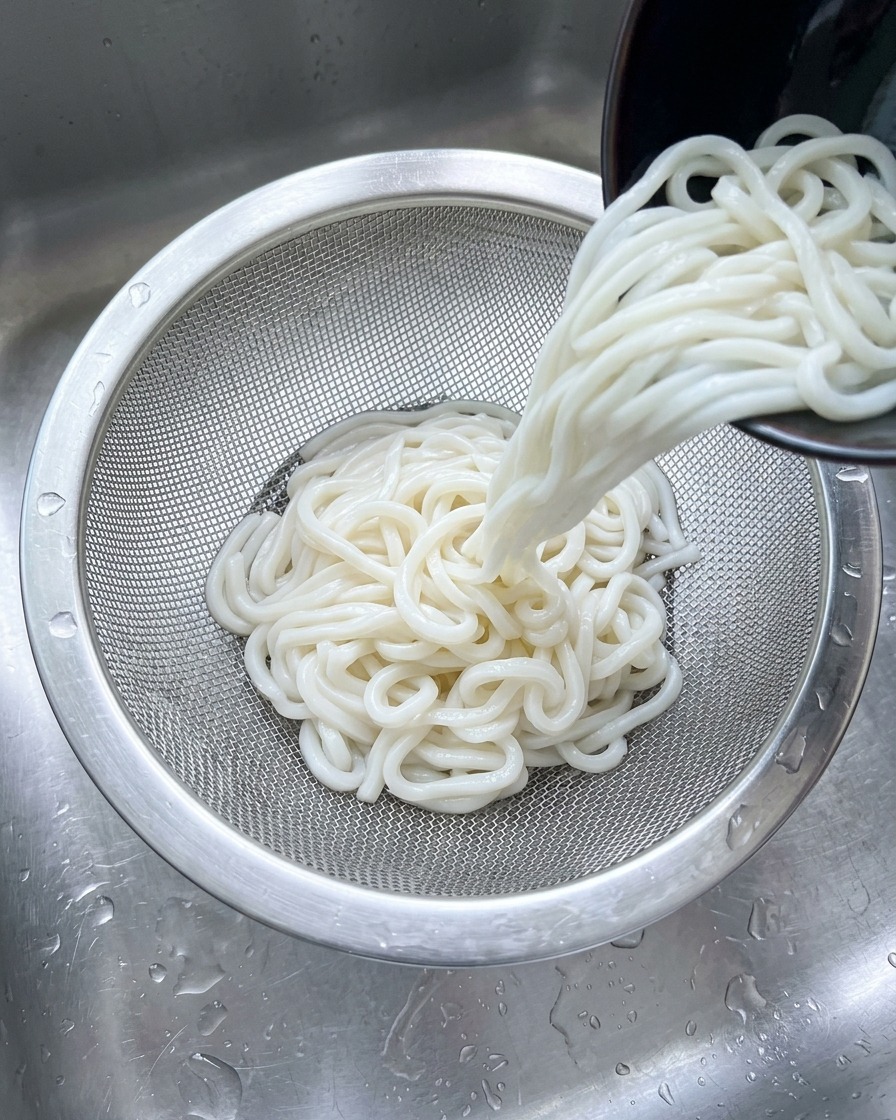Biały makaron udon przesypywany do durszlaka ze stali nierdzewnej pod strumieniem zimnej wody w zlewie.