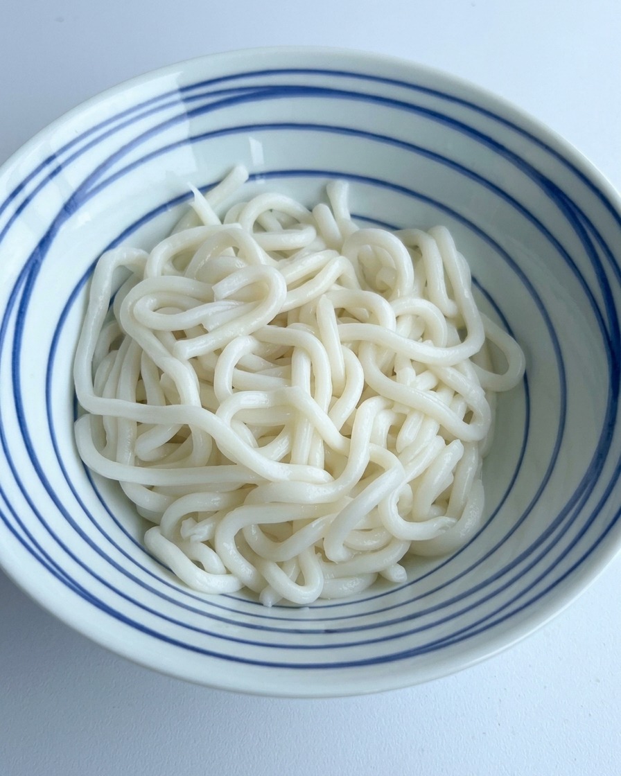 Ciemny sos sojowy i przyprawy wlewane na porcję białego makaronu udon w misce w niebieskie paski.