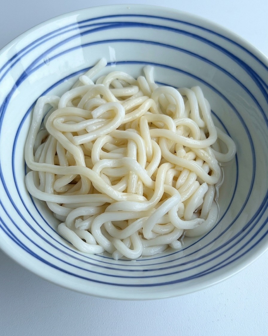 Czysta woda wlewana do miski z makaronem udon, mieszająca się z ciemnymi przyprawami na dnie.