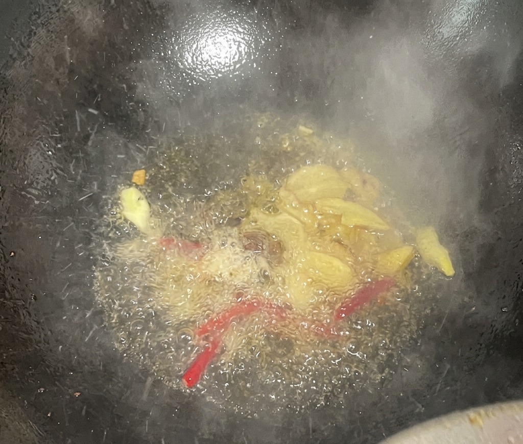 Plasterki imbiru i suszone czerwone papryczki chili skwierczące w gorącym oleju w ciemnym woku, unosząca się para i aromaty.