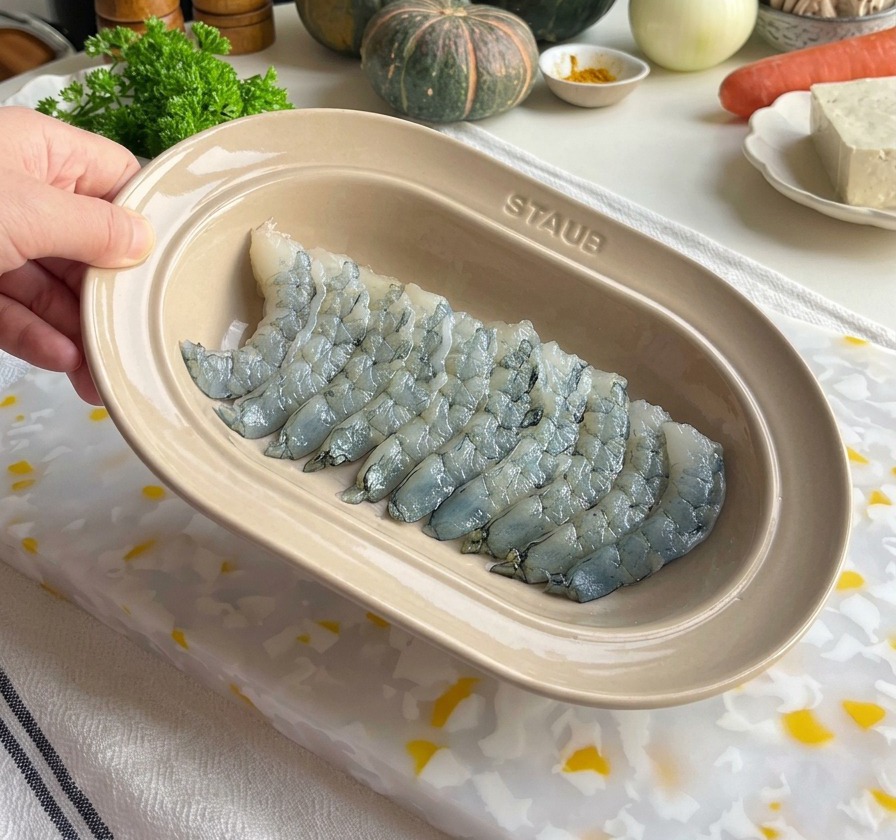 Świeże, oczyszczone surowe krewetki doprawiane w owalnym ceramicznym naczyniu, w tle widoczne młynki do soli i pieprzu.