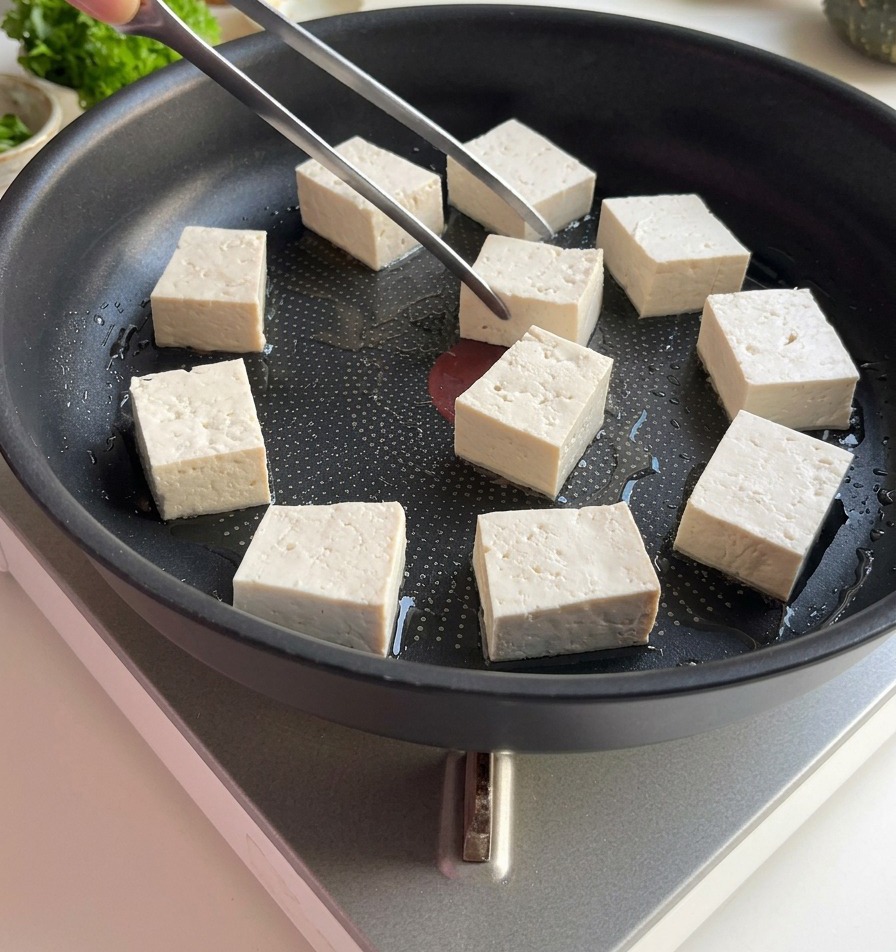 Pęseta układająca białe, kwadratowe kostki tofu na płaskiej patelni z cienką warstwą oleju.