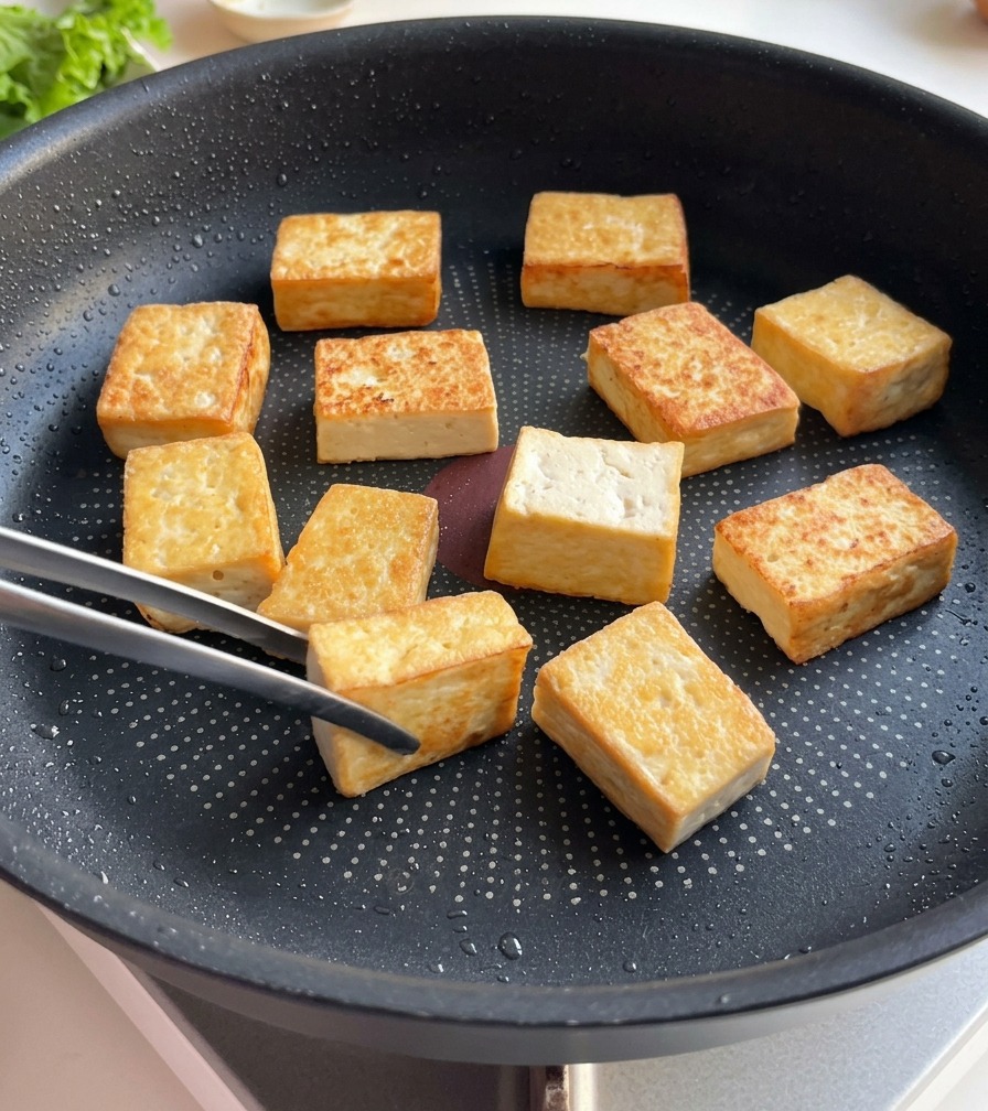 Kostki tofu na patelni z wyraźnie widoczną, głęboką złocistobrązową skórką na usmażonych stronach.