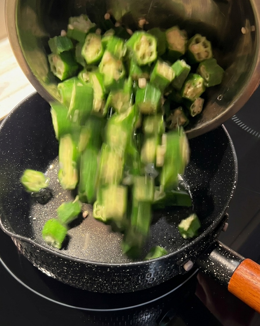 Pokrojona zielona okra wsypywana z metalowej miski na czarną patelnię.
