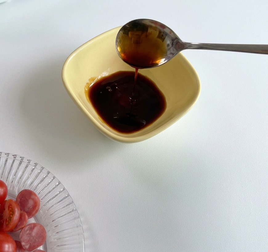 Metalowa łyżka wlewająca ciemny sos sojowy do żółtej miseczki, w której znajduje się już porcja czerwonego keczupu.