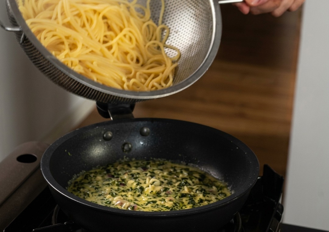 Ugotowane spaghetti przekładane ze stalowego durszlaka na patelnię z kremowym sosem z dodatkiem bazylii.