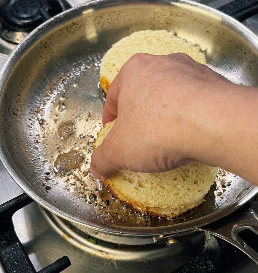 Dłoń dociskająca dwie grube kromki pieczywa brioche na patelni, by uzyskać złocistą skórkę.