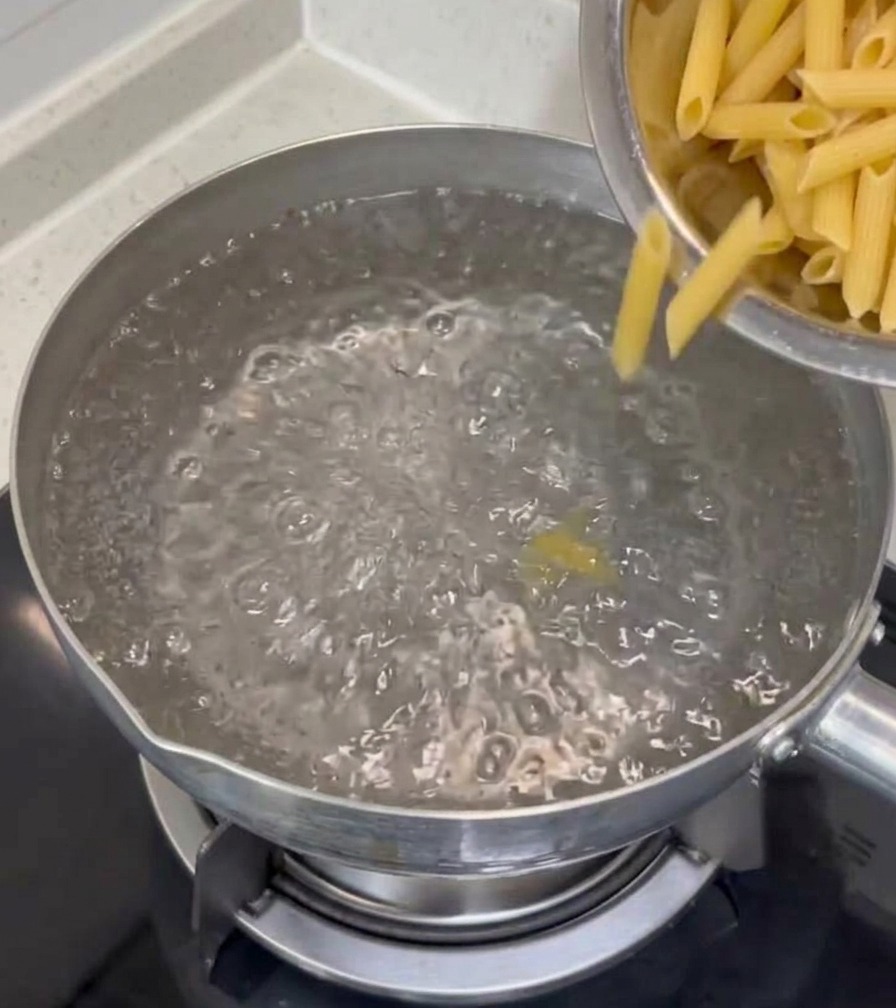 Suchy makaron penne wsypywany z metalowej miski do garnka z bulgoczącą, wrzącą wodą na płycie kuchennej.