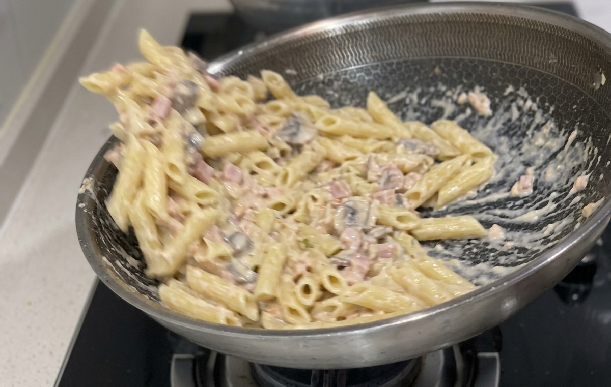 Ugotowany makaron penne mieszany i łączony z kremowym białym sosem i kawałkami łososia na dużej patelni ze stali nierdzewnej.