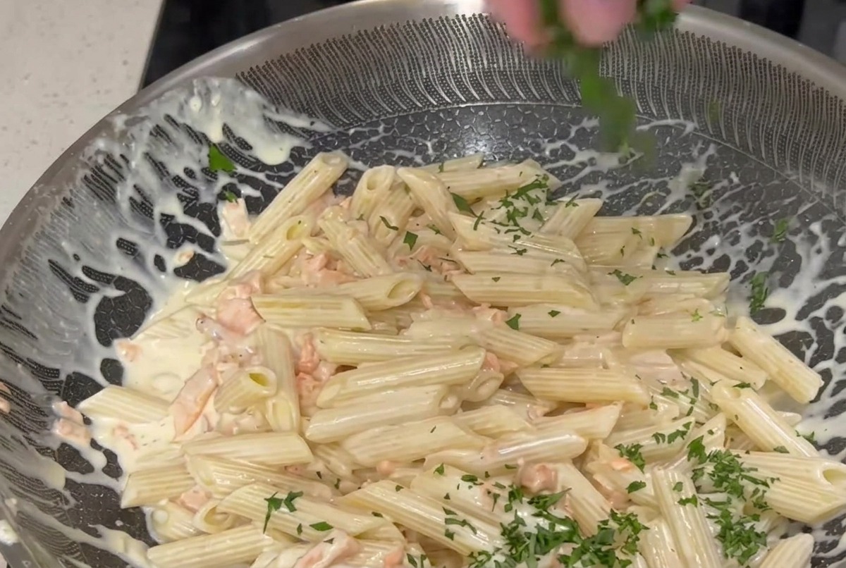 Dłoń posypująca drobno posiekaną zieloną pietruszką dużą patelnię z makaronem penne wymieszanym w kremowym białym sosie z różowymi kawałkami wędzonego łososia.