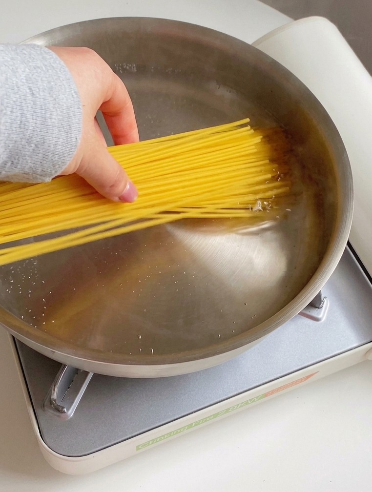 Dłoń wkładająca pęczek suchego makaronu spaghetti do garnka z gotującą się wodą, kroplami oliwy i solą.