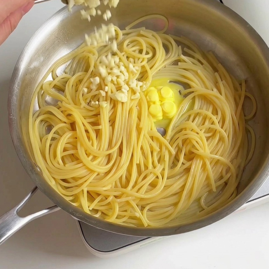 Świeżo posiekany czosnek sypany z łyżki na ugotowane spaghetti i rozpuszczające się masło na patelni.