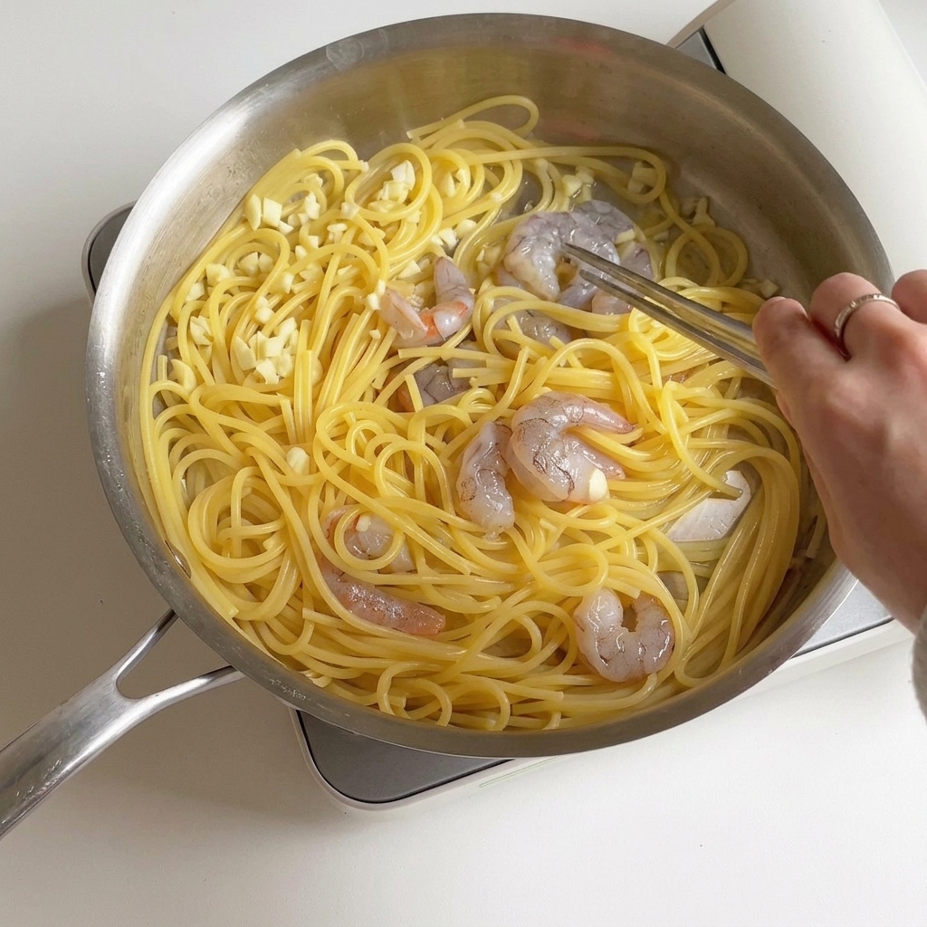 Osoba mieszająca krewetki i spaghetti na patelni, podczas gdy krewetki zmieniają kolor na różowy i matowy.