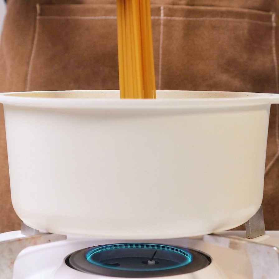 Osoba trzymająca pęczek suchego makaronu spaghetti i wrzucająca go do białego garnka z wrzącą wodą na kuchence gazowej.